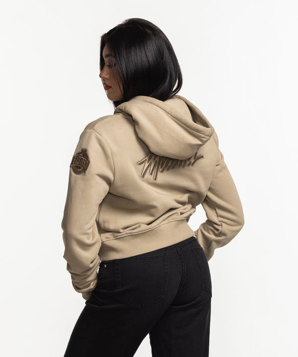 A woman with long dark hair smiles while wearing the Pro Standard Miami HEAT Original Vice Nights Beige Women's Hoodie over a white top and black pants, standing against a plain white background.