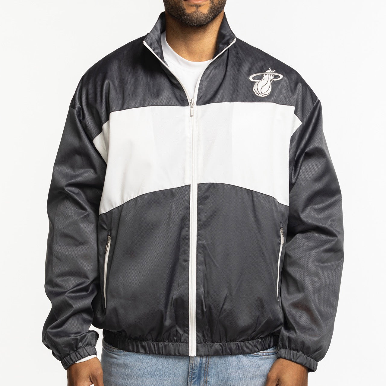 A person wears a Sportiqe Miami HEAT Basketball Club Jacket, featuring a standard fit, black and white zip-up style with team logo on the chest, two front pockets, elastic cuffs, and machine washable polyester; standing against a plain white background.