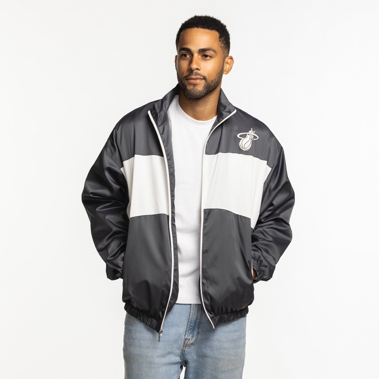 A man wearing a Sportiqe Miami HEAT Basketball Club Jacket by Sportiqe in black and white over a white T-shirt and light blue jeans stands with his hands in his pockets against a plain white background. Machine wash cold for best care.