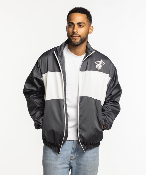 A man wearing a Sportiqe Miami HEAT Basketball Club Jacket by Sportiqe in black and white over a white T-shirt and light blue jeans stands with his hands in his pockets against a plain white background. Machine wash cold for best care.