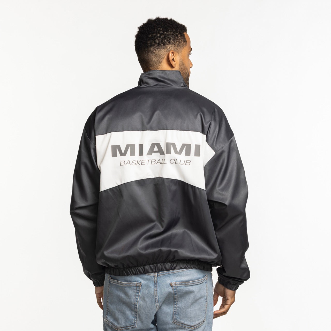 A man with short curly hair wears the Sportiqe Miami HEAT Basketball Club Jacket—a standard fit black and gray polyester piece by Sportiqe with 