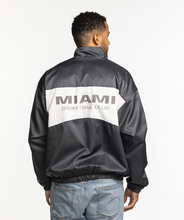 A man wearing a Sportiqe Miami HEAT Basketball Club Jacket by Sportiqe in black and white over a white T-shirt and light blue jeans stands with his hands in his pockets against a plain white background. Machine wash cold for best care.