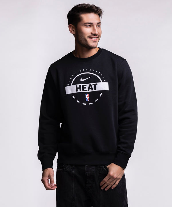 A man smiles and looks left while wearing the Nike Miami HEAT 2025-26 On-Court Practice Crewneck, featuring the Nike logo and HEAT text on the front, standing against a plain light background.