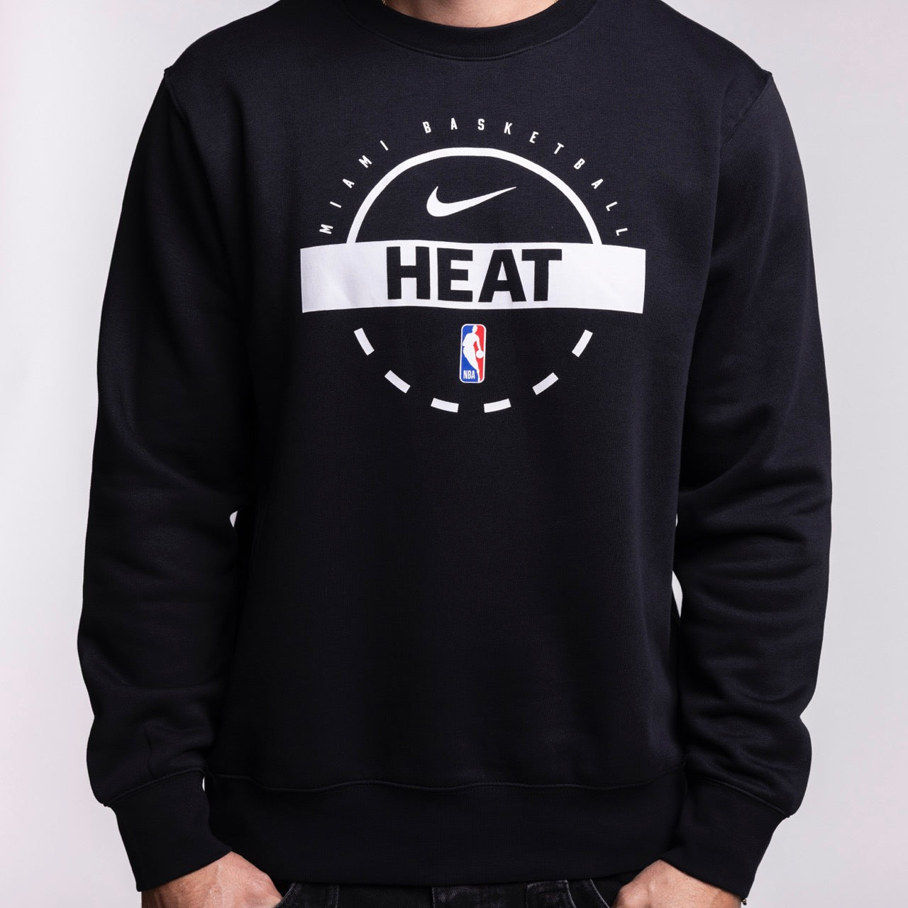 Someone wears the Nike Miami HEAT 2025-26 On-Court Practice Crewneck, a classic fit black sweatshirt with Nike and NBA logos, HEAT and Miami Basketball text, cozy fleece lining, and hands in pockets, set against a plain background.