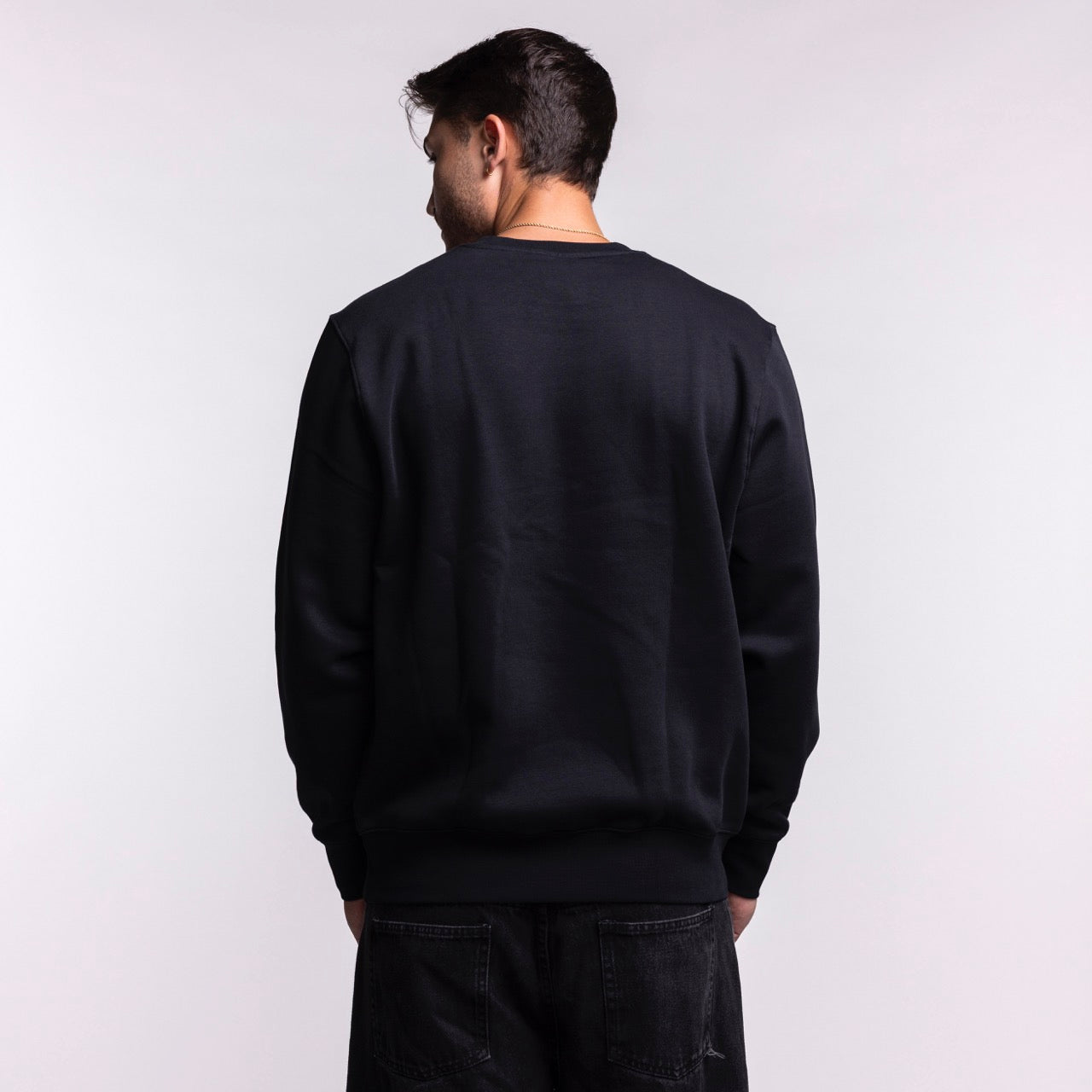 A man with short dark hair stands facing away in a Nike Miami HEAT 2025-26 On-Court Practice Crewneck and black jeans, set against a light background.