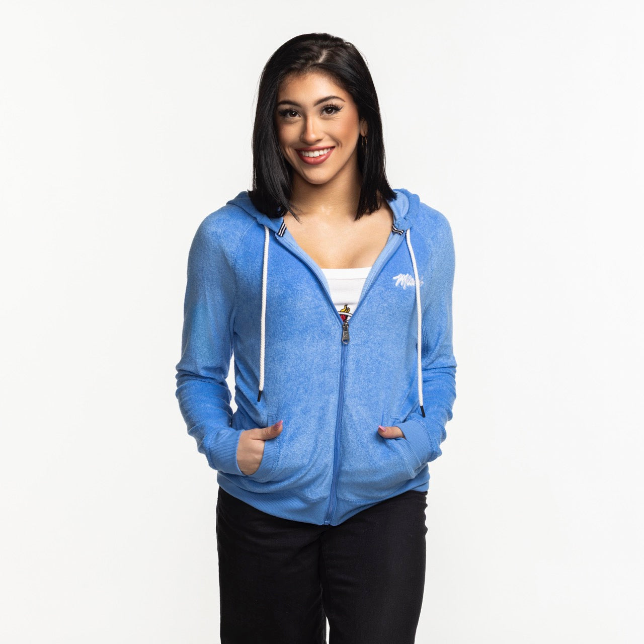 A woman with straight, shoulder-length dark hair smiles at the camera wearing the Antigua Miami HEAT Original Vice Nights Women's Zip Up Jacket by Antigua over a white top, hands in pockets, standing against a plain white background.