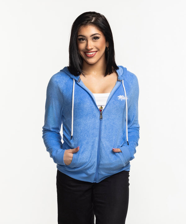 A woman with straight, shoulder-length dark hair smiles at the camera wearing the Antigua Miami HEAT Original Vice Nights Women's Zip Up Jacket by Antigua over a white top, hands in pockets, standing against a plain white background.