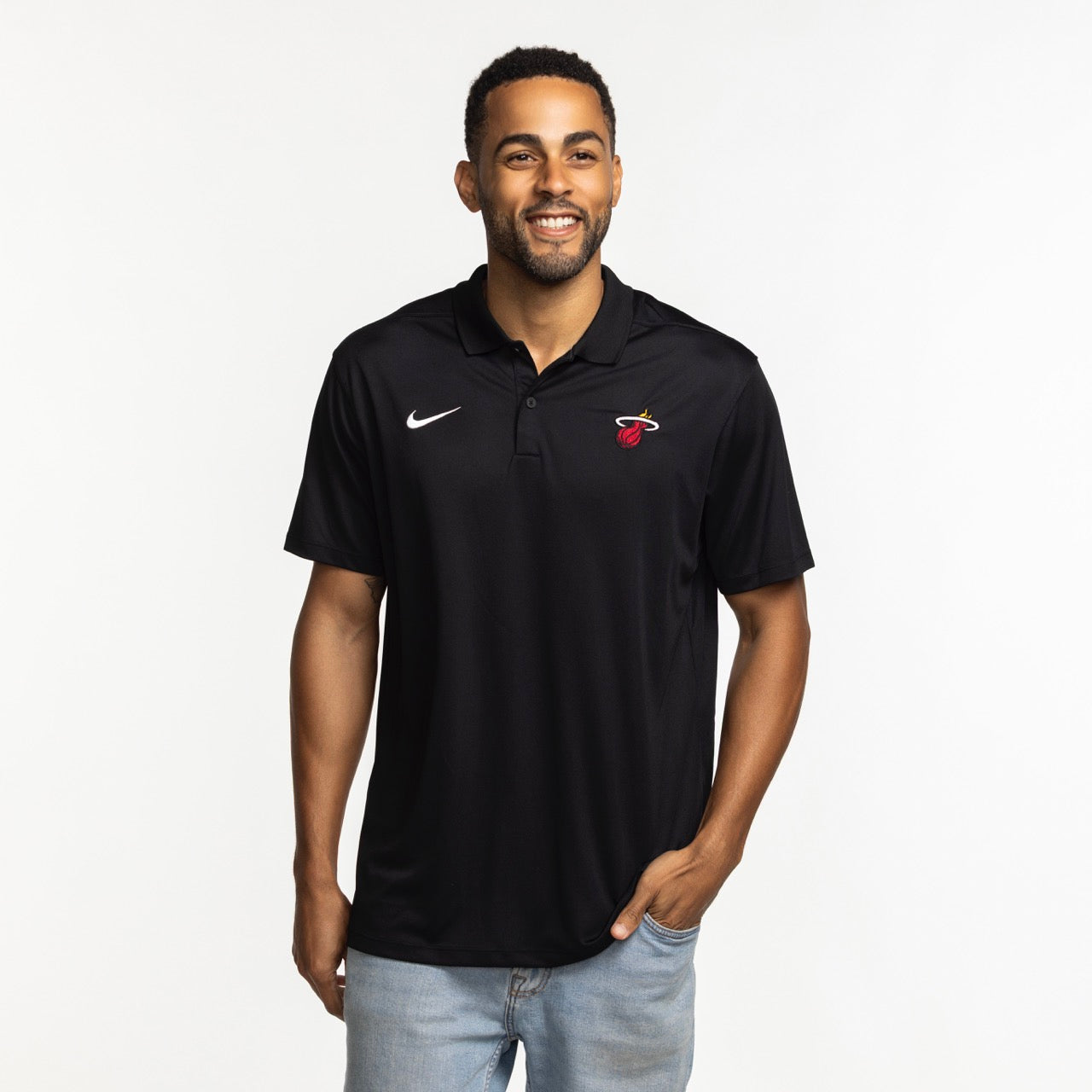 A smiling man wears the Nike Miami HEAT Logo Victory Polo—a black polyester shirt with a white Nike swoosh and Miami Heat logo, standard fit—standing against a plain white background with one hand in his pocket.
