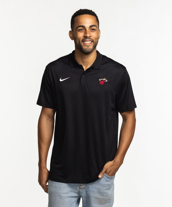 A smiling man wears the Nike Miami HEAT Logo Victory Polo—a black polyester shirt with a white Nike swoosh and Miami Heat logo, standard fit—standing against a plain white background with one hand in his pocket.
