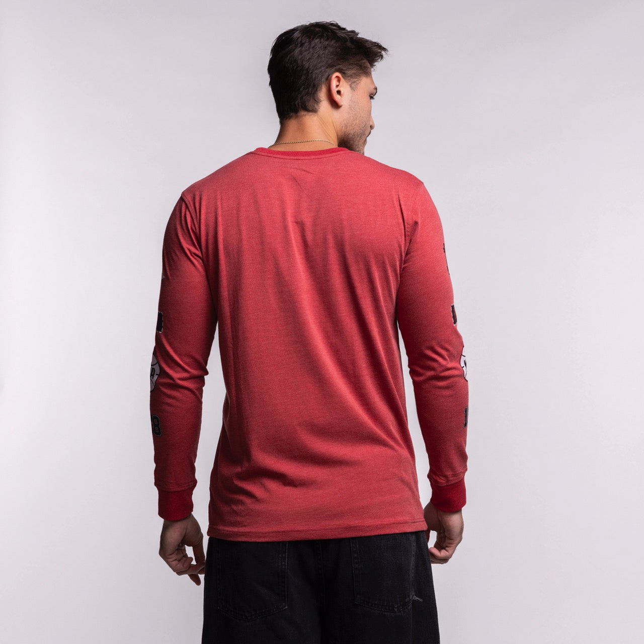 A man stands with his back to the camera wearing a New Era Miami HEAT Red Cuffed Long Sleeve Tee and black pants against a plain light background, his arms relaxed at his sides.