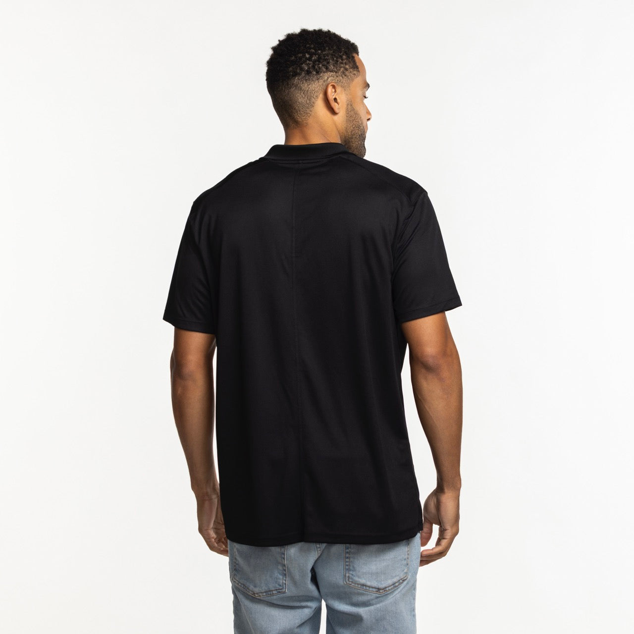 A man faces away from the camera, wearing a black Nike Miami HEAT Logo Victory Polo and light blue jeans against a plain white background.