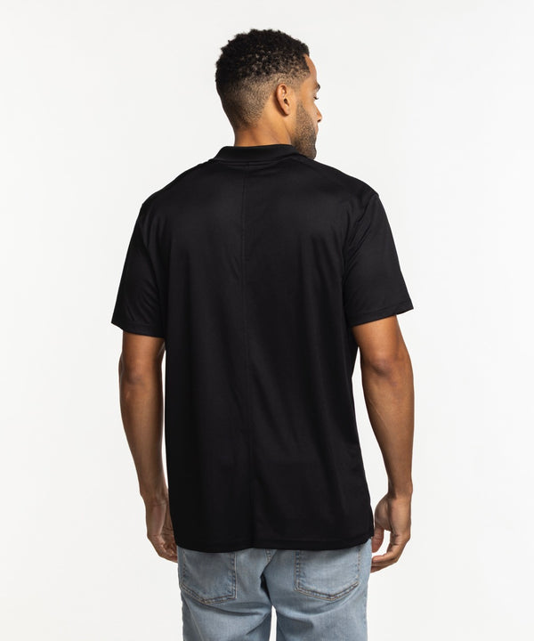 A smiling man wears the Nike Miami HEAT Logo Victory Polo—a black polyester shirt with a white Nike swoosh and Miami Heat logo, standard fit—standing against a plain white background with one hand in his pocket.