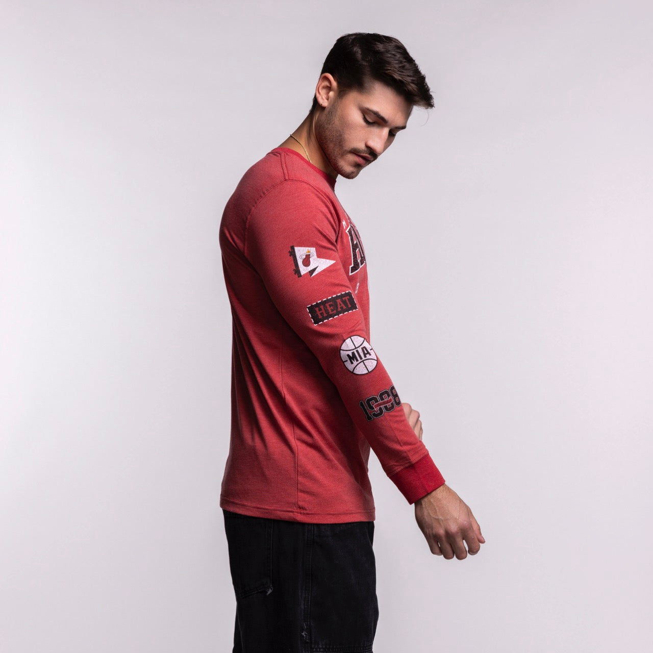 A man wearing the New Era Miami HEAT Red Cuffed Long Sleeve Tee stands sideways against a plain white background, looking down while adjusting his sleeve. Machine wash cold for best care.