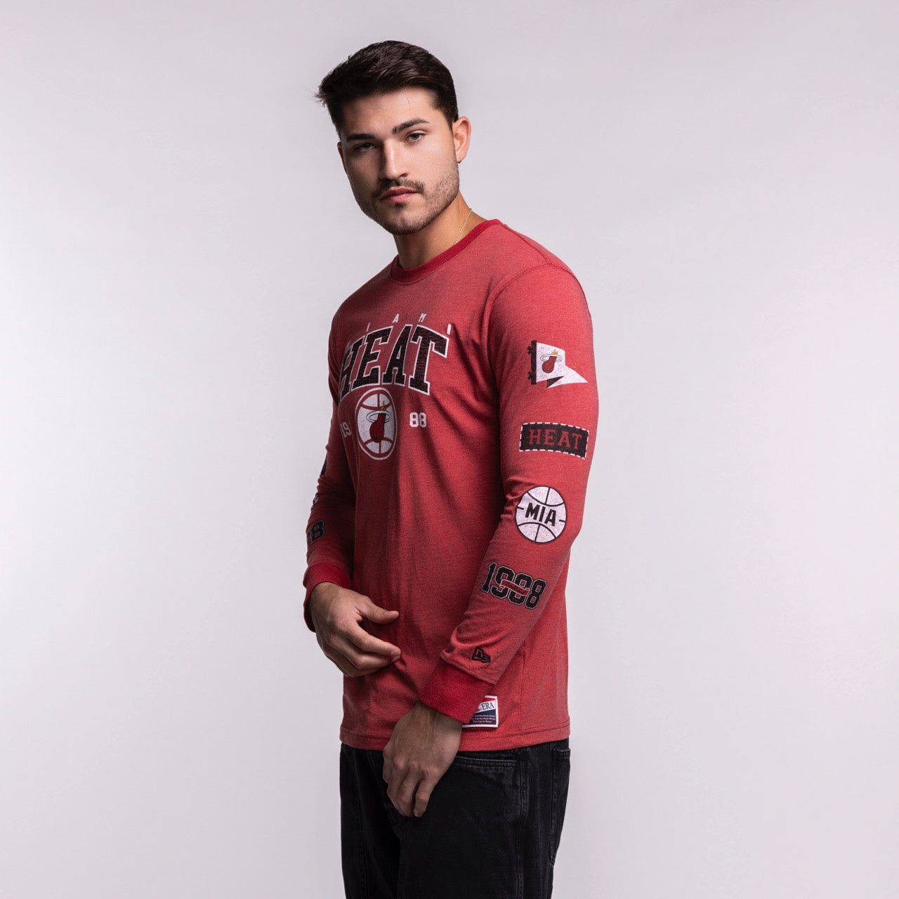 A man wears the New Era Miami HEAT Red Cuffed Long Sleeve Tee, featuring team logos and patches, made from a cotton-polyester blend for comfort and durability, standing against a light gray background with a neutral expression.