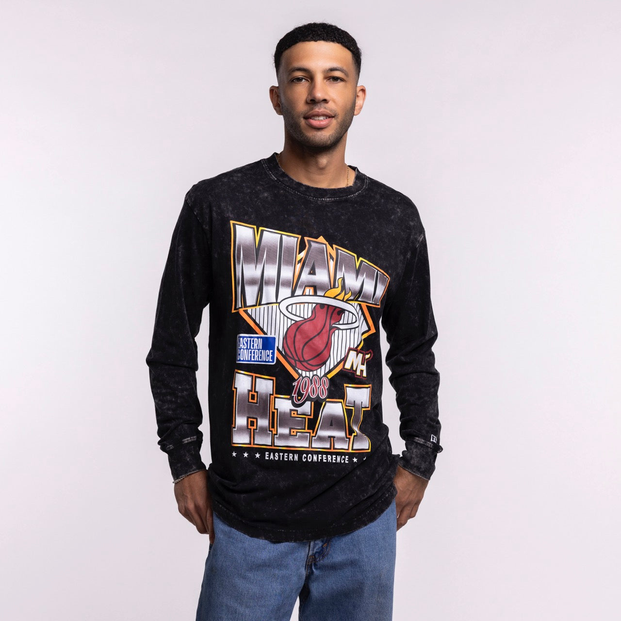 A man faces forward against a plain light background, wearing blue jeans and the New Era Miami HEAT Washed Long Sleeve Tee—a black, oversized shirt with bold graphics from New Era.