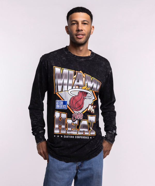 A man faces forward against a plain light background, wearing blue jeans and the New Era Miami HEAT Washed Long Sleeve Tee—a black, oversized shirt with bold graphics from New Era.