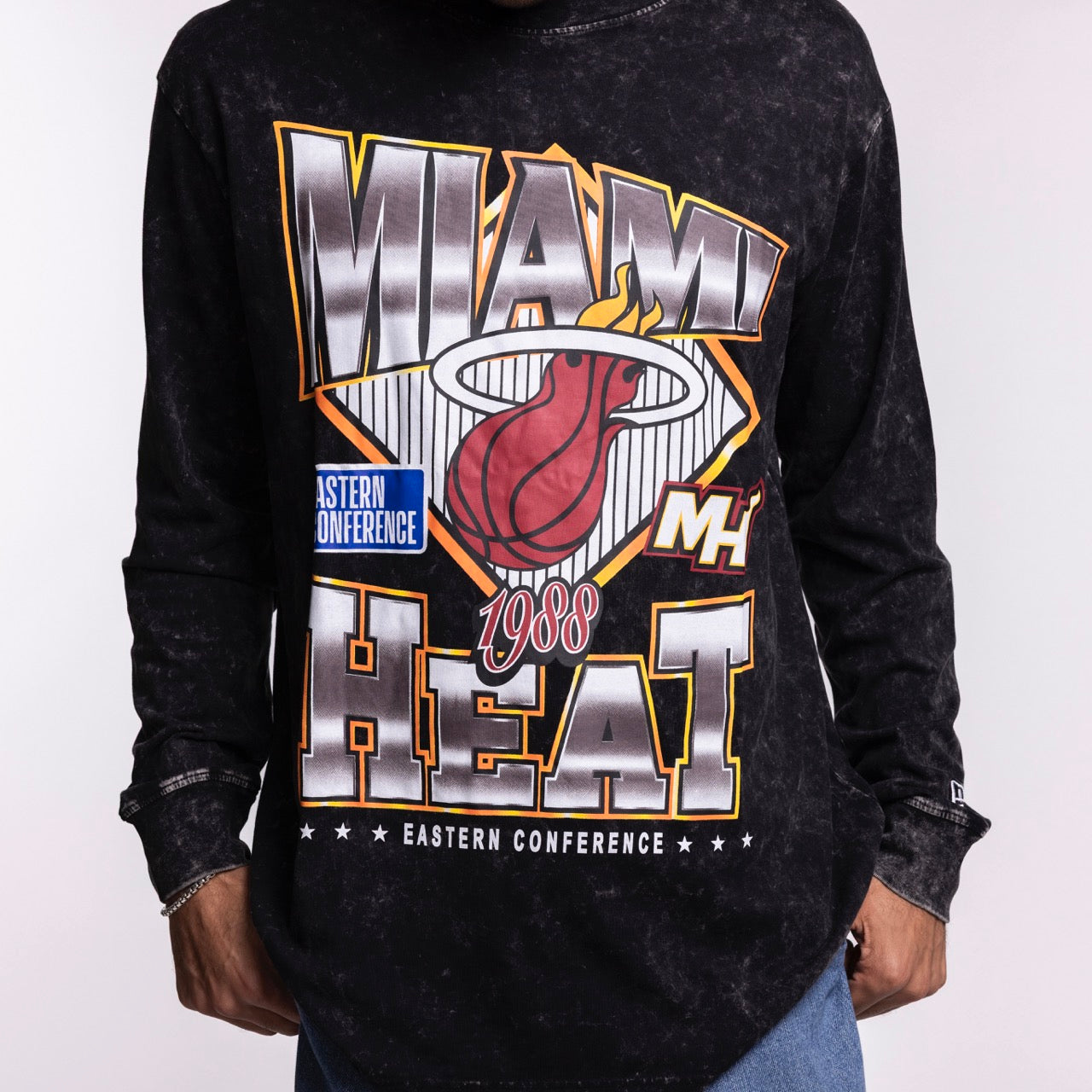 A person wearing the New Era Miami HEAT Washed Long Sleeve Tee—a black, oversized 100% cotton shirt featuring a colorful Miami Heat graphic with the team logo, 