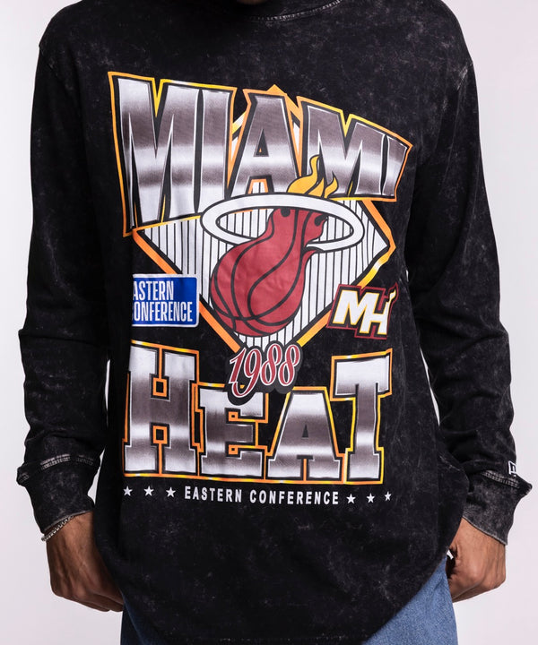 A man faces forward against a plain light background, wearing blue jeans and the New Era Miami HEAT Washed Long Sleeve Tee—a black, oversized shirt with bold graphics from New Era.
