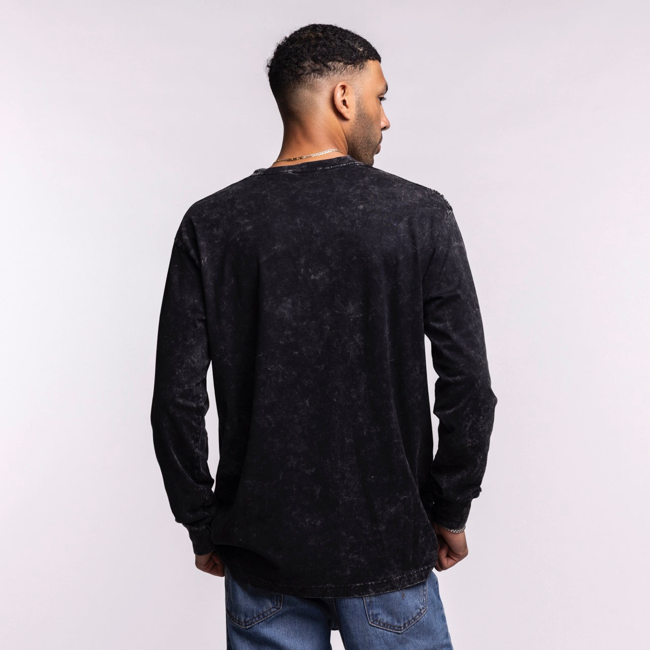 A man faces away from the camera, wearing the New Era Miami HEAT Washed Long Sleeve Tee in black with an oversized fit and blue jeans, against a plain light background.