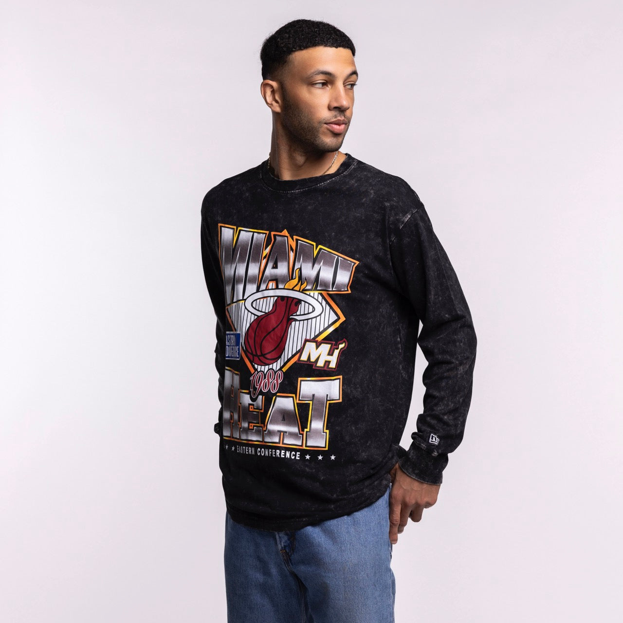A man wears the New Era Miami HEAT Washed Long Sleeve Tee, featuring colorful graphics and a washed pattern, as he stands against a plain light background looking left with one hand by his side and the other on his hip.