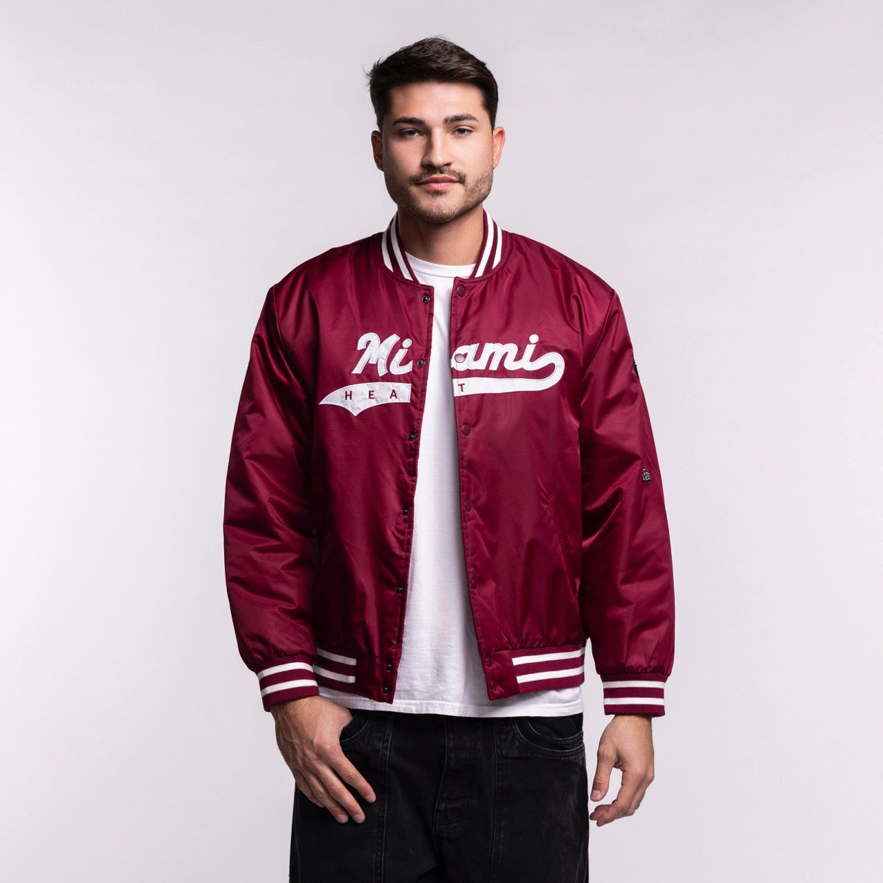 A man faces forward wearing a Hype and Vice Miami HEAT Varsity Jacket in maroon with white script, paired with a white t-shirt and black pants, set against a plain light background.