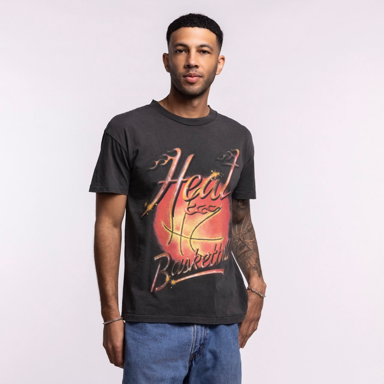 A man with short hair and an arm tattoo stands against a light background in blue jeans, wearing the Court Culture 