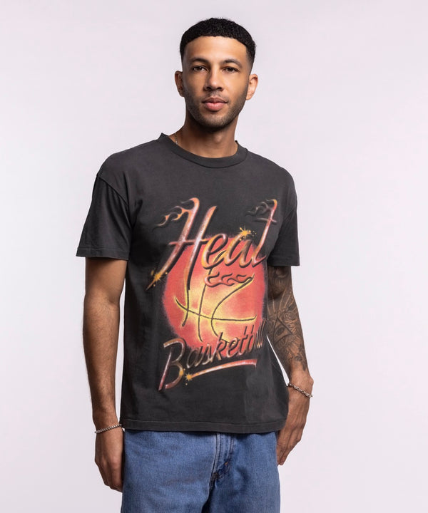 A man with short hair and an arm tattoo stands against a light background in blue jeans, wearing the Court Culture 