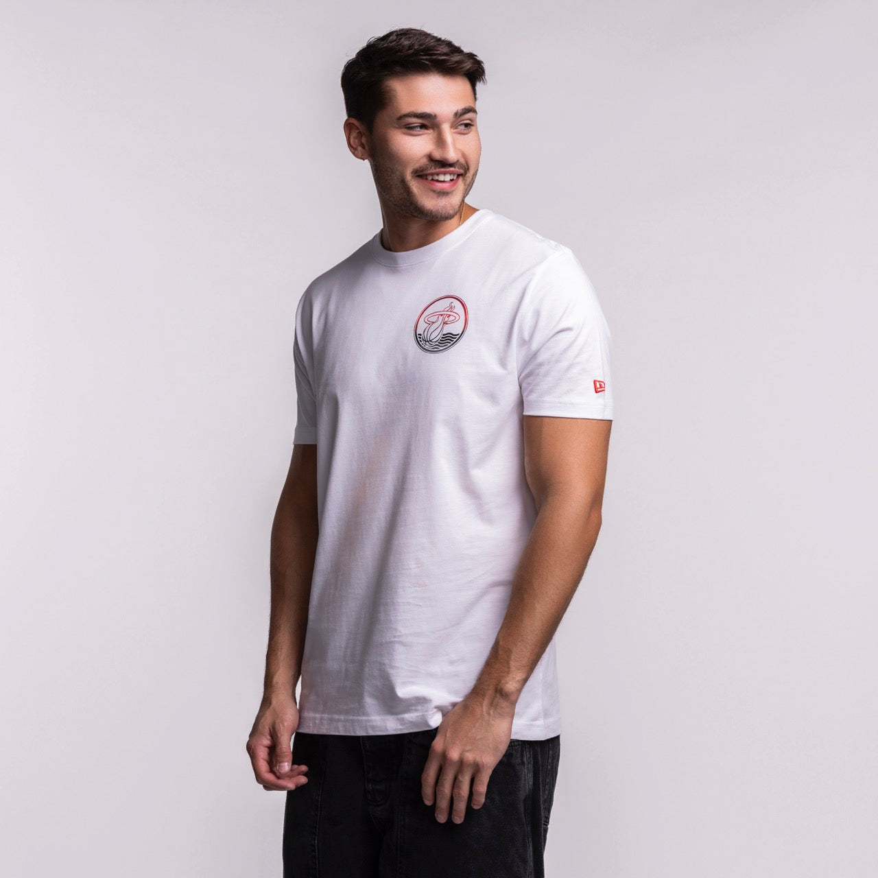 A man with short dark hair and a beard stands sideways, smiling in a New Era Miami HEAT White Basketball Tee and black pants, against a plain light background.