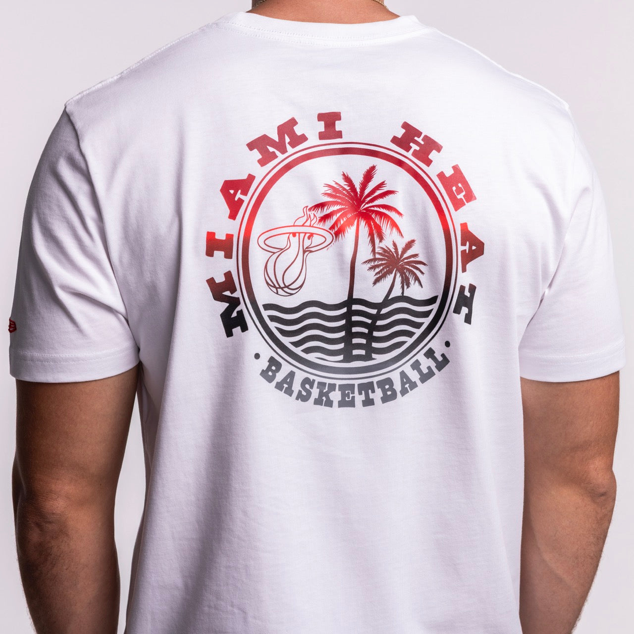 A person wears the New Era Miami HEAT White Basketball Tee, a 100% cotton, standard-fit T-shirt with Miami Heat logo, red palm trees, ocean waves, and 
