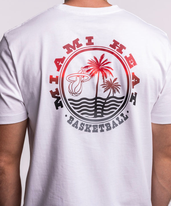 A man with short dark hair and a beard stands sideways, smiling in a New Era Miami HEAT White Basketball Tee and black pants, against a plain light background.