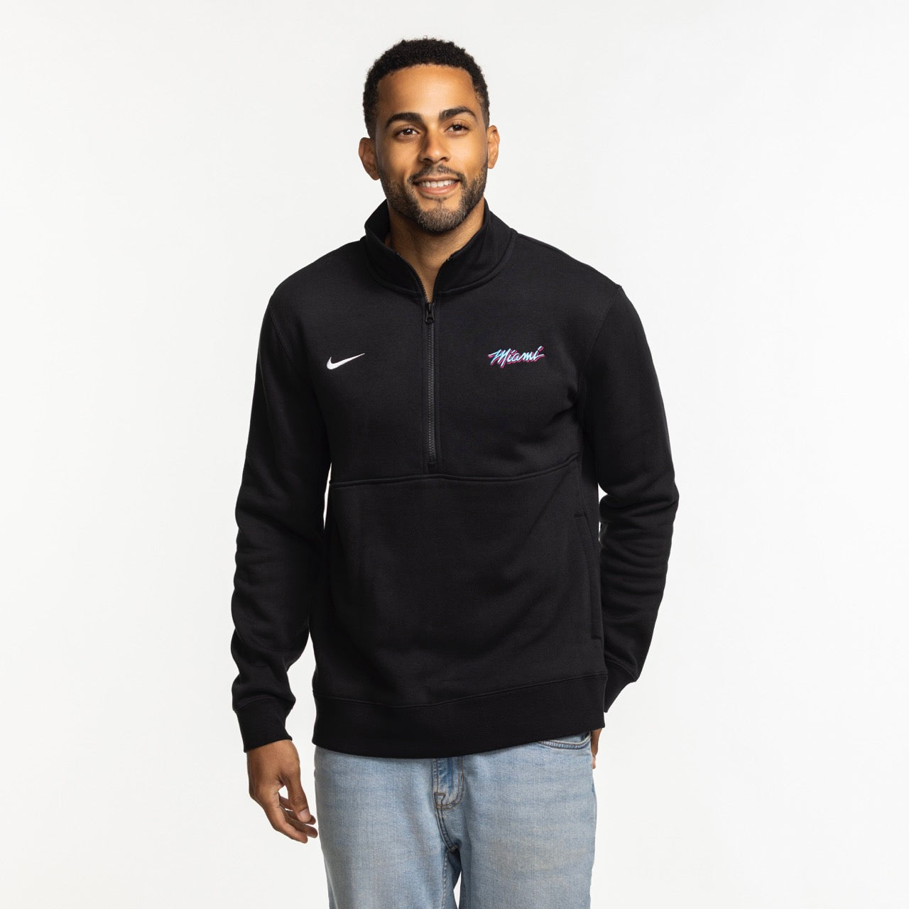 A man smiles against a white background wearing the Nike Miami HEAT Original Vice Nights 1/2 Zip Fleece Jacket—standard fit, made from a cotton-poly blend, featuring “Miami” on the chest—paired with light blue jeans.