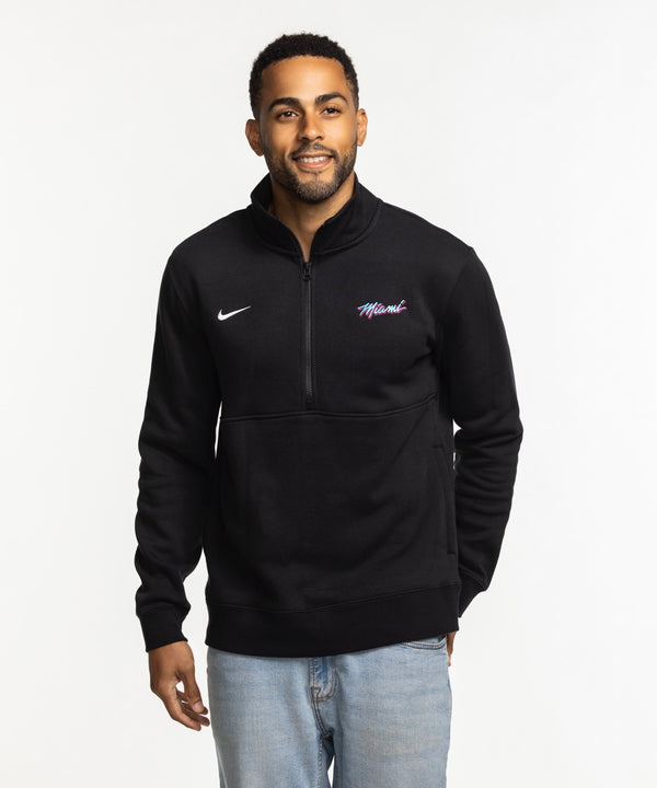 A man smiles against a white background wearing the Nike Miami HEAT Original Vice Nights 1/2 Zip Fleece Jacket—standard fit, made from a cotton-poly blend, featuring “Miami” on the chest—paired with light blue jeans.