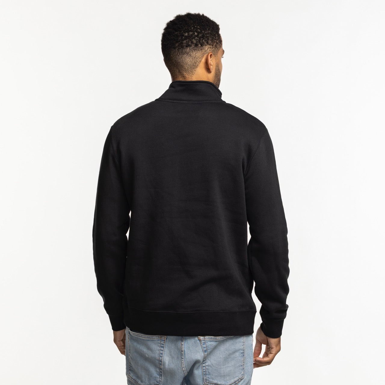 A man with short curly hair, seen from behind, wears a Nike Miami HEAT Original Vice Nights 1/2 Zip Fleece Jacket and light blue jeans, standing against a plain white background.