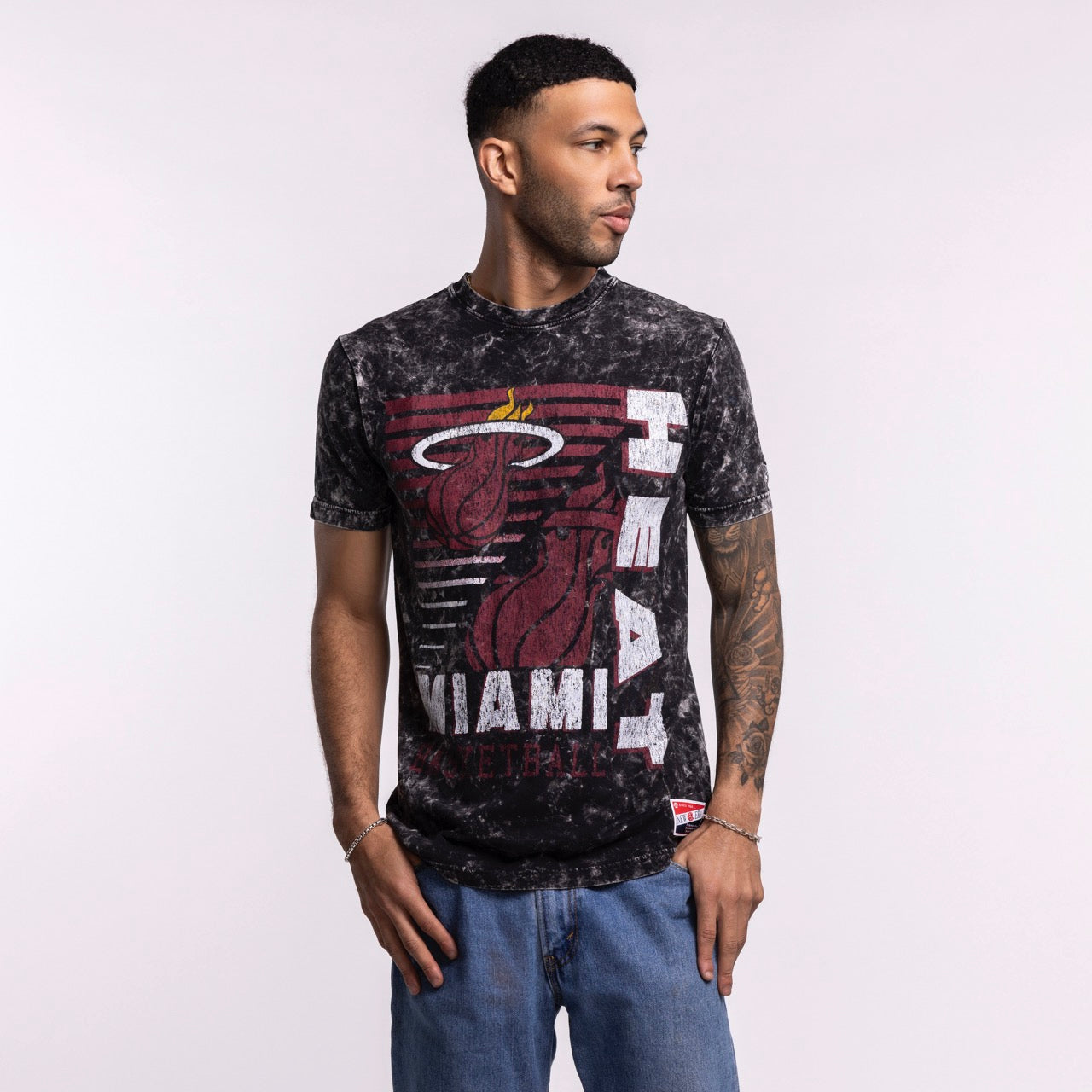 A man wears the New Era Miami HEAT Mineral Washed Tee, a black and gray 100% cotton shirt with a large team logo, standing against a light background and looking to his left with one hand in his pocket.