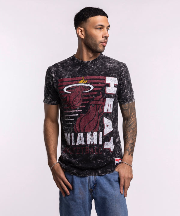A man wears the New Era Miami HEAT Mineral Washed Tee, a black and gray 100% cotton shirt with a large team logo, standing against a light background and looking to his left with one hand in his pocket.