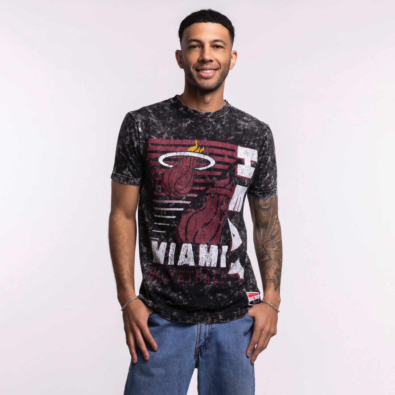 A smiling man with short hair and a tattoo on his left arm wears the New Era Miami HEAT Mineral Washed Tee and blue jeans, standing against a plain light background. Machine wash cold.