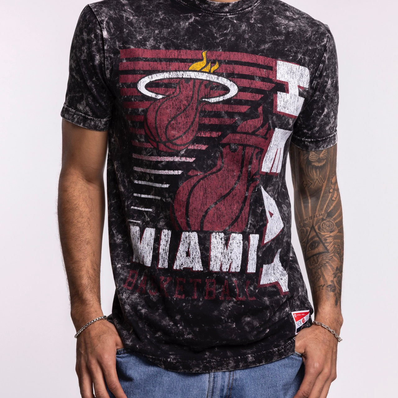 A person wears the New Era Miami HEAT Mineral Washed Tee—a black and white tie-dye shirt by New Era with the team's logo and name in bold red and white. Only the upper body is visible; one arm sports a tattoo sleeve.