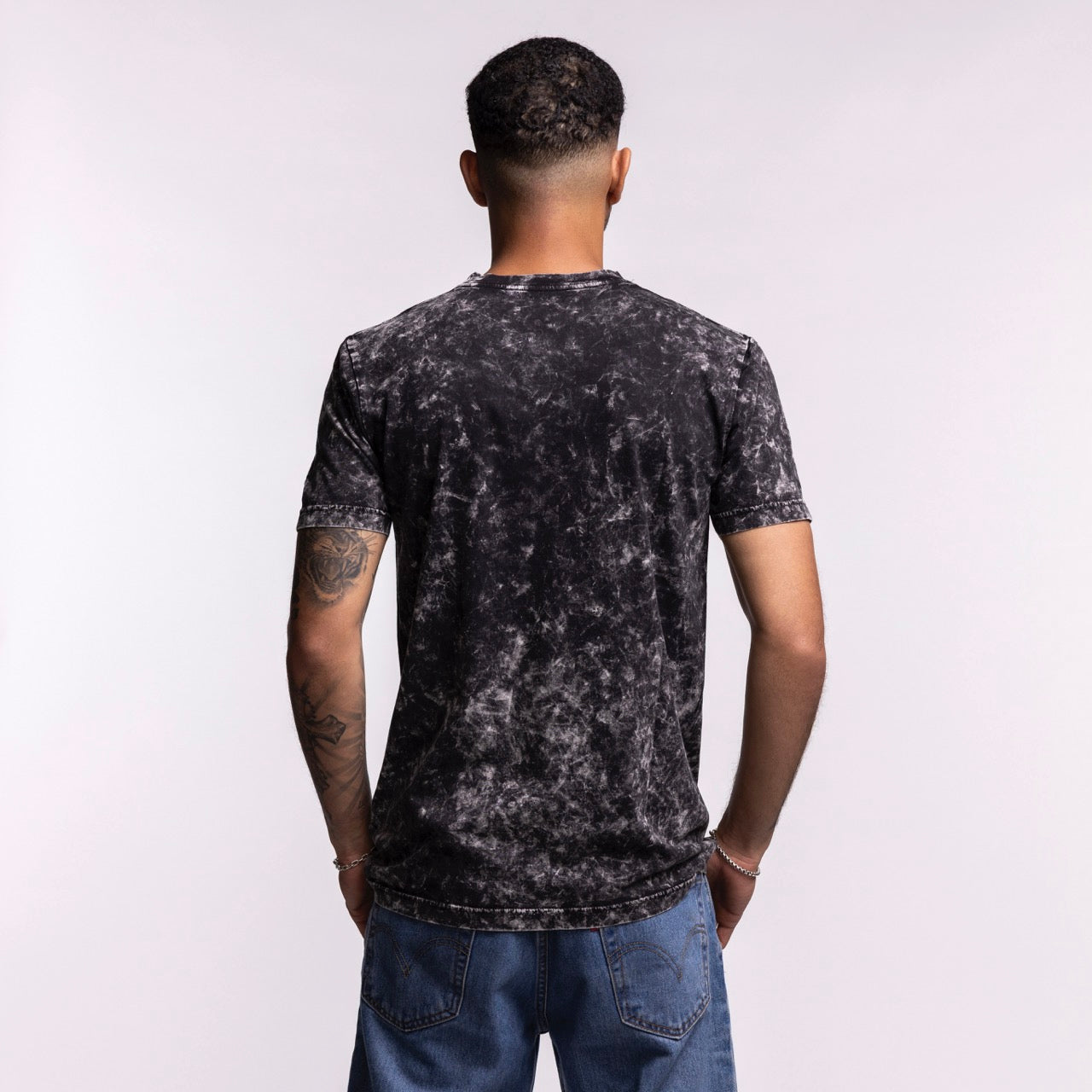 A person with short hair, seen from behind, wears the New Era Miami HEAT Mineral Washed Tee by New Era—a black and gray marbled, 100% cotton standard fit t-shirt—with blue jeans, set against a plain light background.
