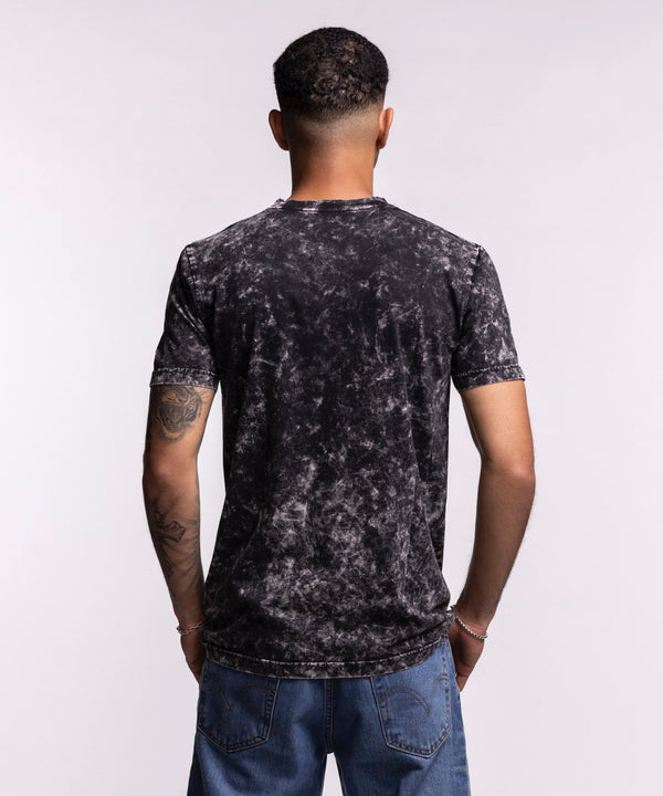 A man wears the New Era Miami HEAT Mineral Washed Tee, a black and gray 100% cotton shirt with a large team logo, standing against a light background and looking to his left with one hand in his pocket.