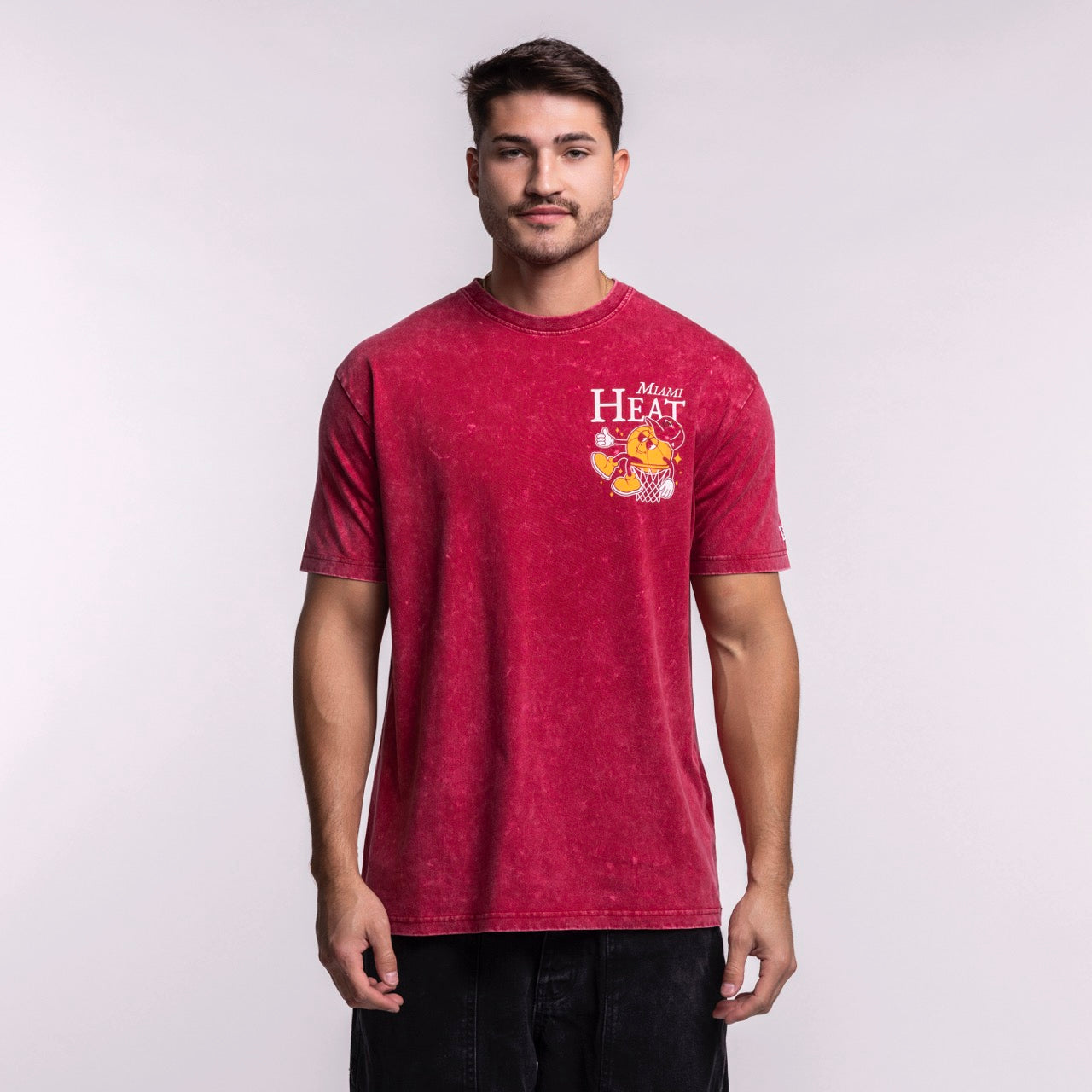 A man wears the New Era Miami HEAT Ball Character Tee, featuring a cartoon mascot graphic on red. This standard fit t-shirt by New Era is made of 100% cotton and is machine washable for easy care, shown against a plain light background.