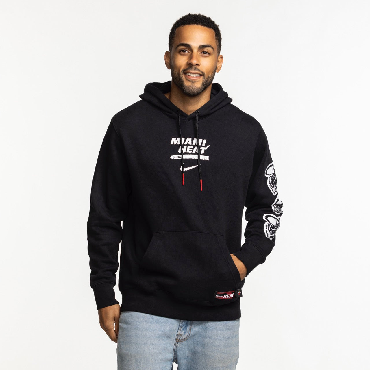 A man stands against a white background, wearing a Nike Miami HEAT Club Pullover Black Hoodie with MIAMI HEAT and a basketball graphic, 100% cotton hood lining, light blue jeans, and a slight smile.