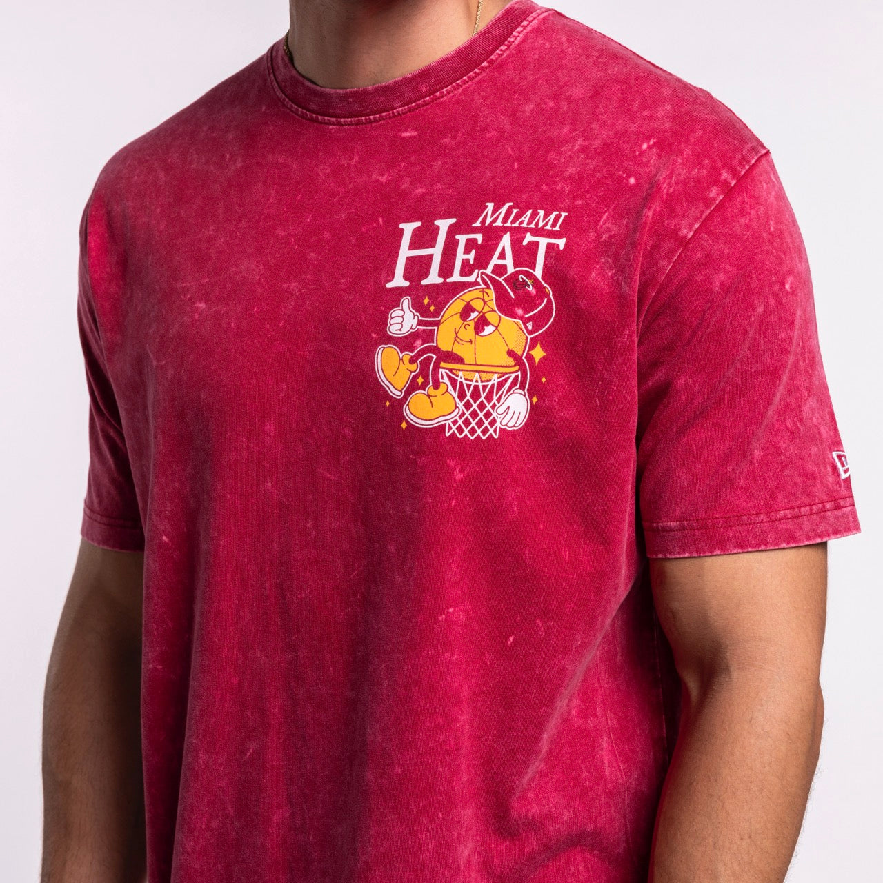 A person is wearing the New Era Miami HEAT Ball Character Tee—a 100% cotton red t-shirt by New Era featuring a cartoon basketball character dunking, with the team name and logo printed on the chest. Standard fit.