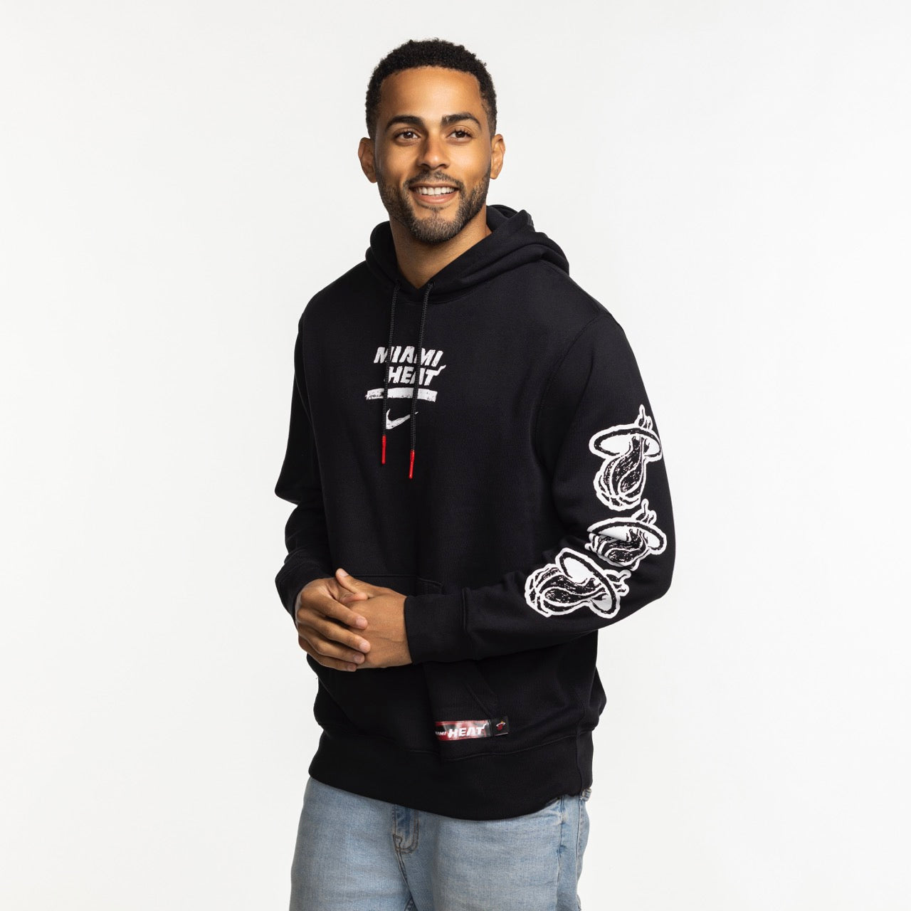 A man stands smiling in a Nike Miami HEAT Club Pullover Black Hoodie featuring team graphics on the chest and sleeve. Made from 100% cotton with a cozy hood lining, he pairs it with light blue jeans against a white background.