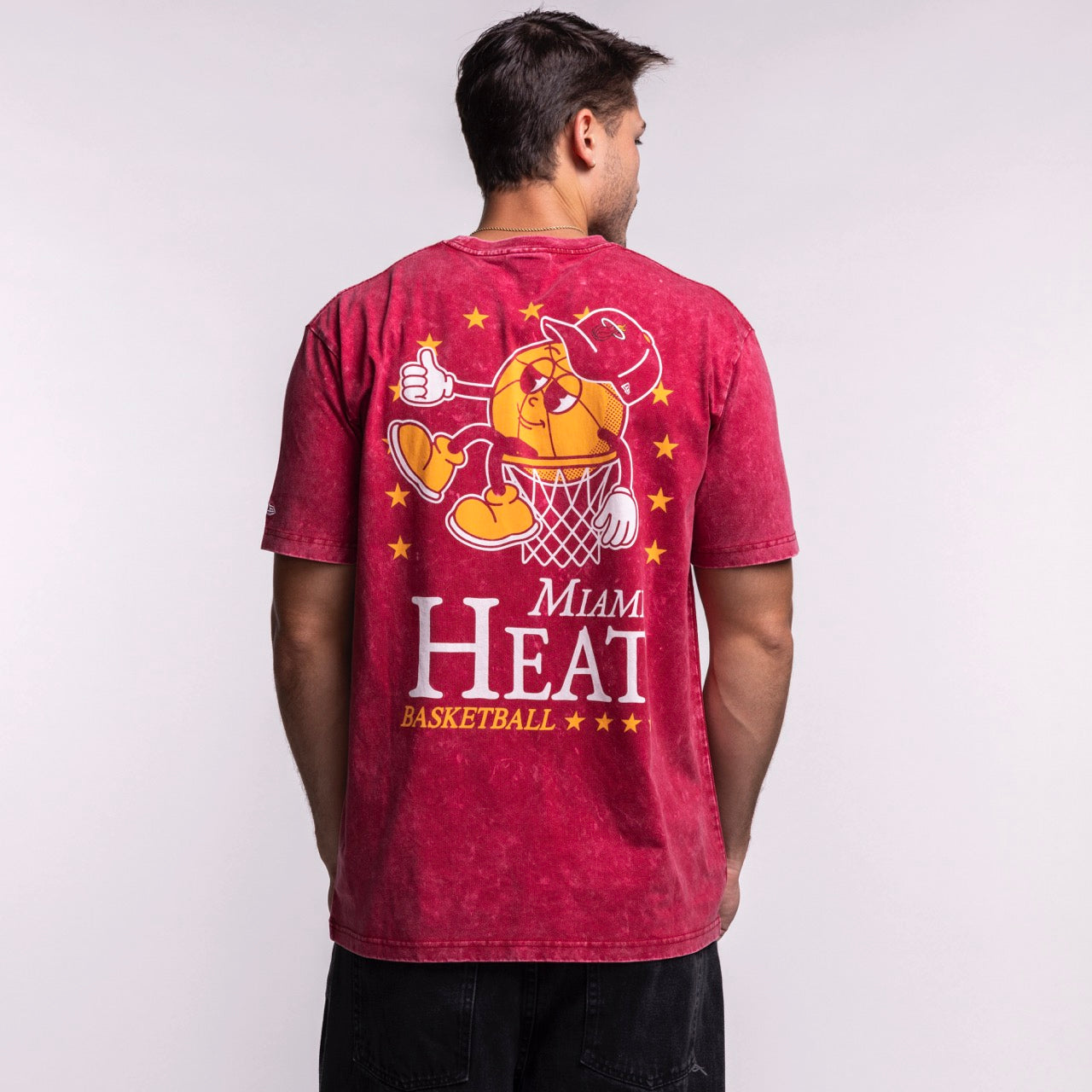 A man faces away wearing the New Era Miami HEAT Ball Character Tee—a 100% cotton, standard fit T-shirt from New Era featuring a cartoon dunk, stars, and text on the back. Easy care: machine wash cold.