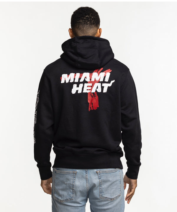 A man stands against a white background, wearing a Nike Miami HEAT Club Pullover Black Hoodie with MIAMI HEAT and a basketball graphic, 100% cotton hood lining, light blue jeans, and a slight smile.