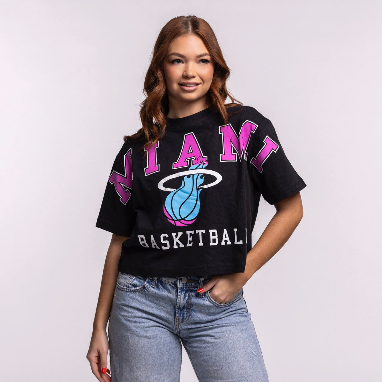 A woman with long brown hair smiles with one hand in her pocket, wearing a relaxed fit black '47 Brand Miami HEAT Original Vice Nights Basketball Women's Tee and blue jeans against a plain light background.