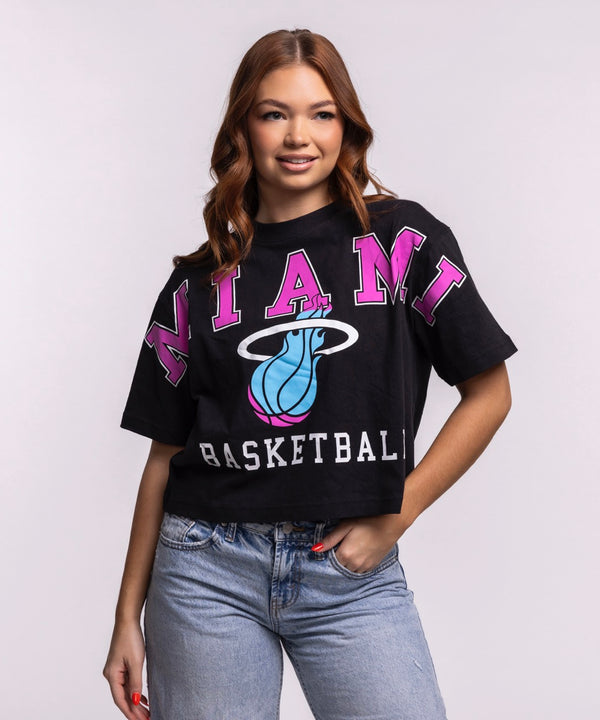 A woman with long brown hair wears the '47 Brand Miami HEAT Original Vice Nights Basketball Women's Tee, a relaxed fit black cropped T-shirt featuring MIAMI, BASKETBALL, and a basketball logo, paired with light blue jeans against a plain background.