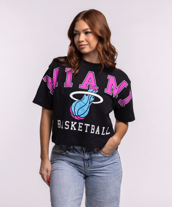 A woman with long brown hair wears the '47 Brand Miami HEAT Original Vice Nights Basketball Women's Tee, a relaxed fit black cropped T-shirt featuring MIAMI, BASKETBALL, and a basketball logo, paired with light blue jeans against a plain background.