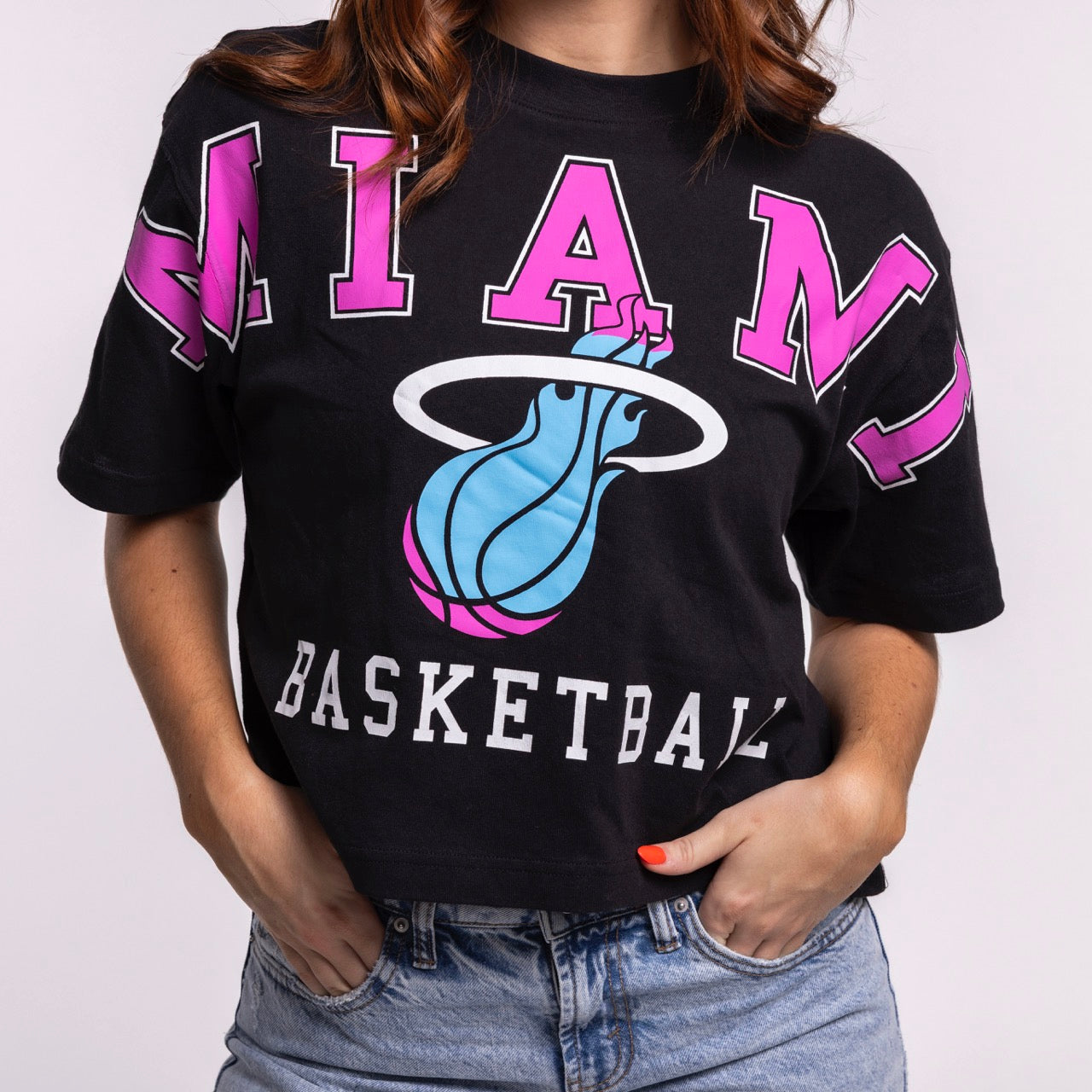 A woman wears the '47 Brand Miami HEAT Original Vice Nights Basketball Women's Tee—a black cropped t-shirt with “MIAMI BASKETBALL” and a flaming basketball logo in pink, blue, and white—paired with light blue denim jeans. 100% cotton; machine wash cold.