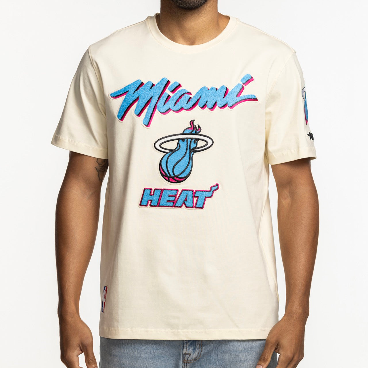 A person wears the Pro Standard Miami HEAT Original Vice Nights Logo Tee—a cream, standard-fit t-shirt with blue and pink Miami Heat graphics—paired with light blue jeans, shown waist up against a plain white background.
